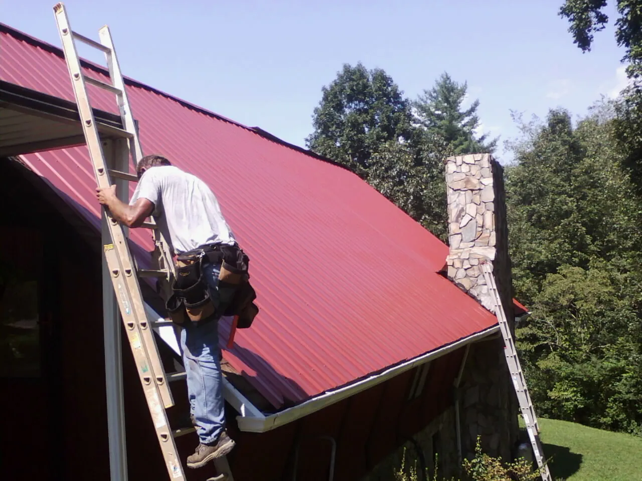 Roofer on a ladder beside a red metal roof and stone chimney — Soffit & Fascia Repair by Roofing Brothers in Saginaw